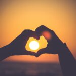duas mãos unidas formando um coração com o por do sol ao centro