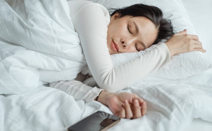 Calm Asian female wearing white pajama sleeping in comfortable bed with white sheets near modern mobile phone in morning