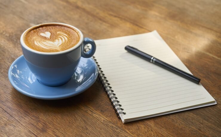 cappuccino em xícara de cerâmica azul, caderno com caneta ao lado