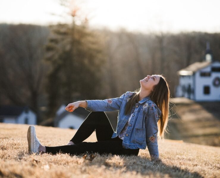 donna felice in giacca di jeans blu seduta sull'erba durante il giorno