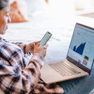 mulher olhando celular na cama com notebook na frente