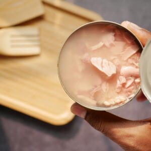 A close-up of an opened canned tuna held over a wooden cutting board and utensils.