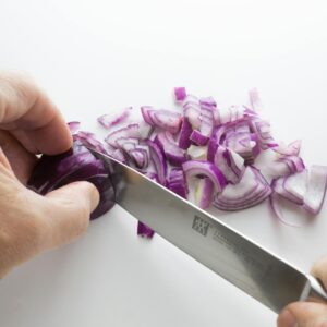 Close-up of hands slicing purple onions on a white cutting board with a sharp knife.