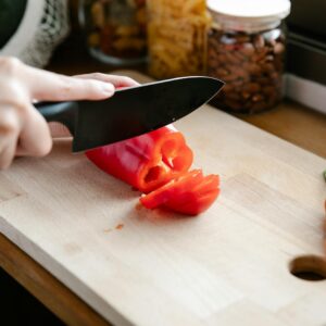 A person slicing fresh red bell pepper on a wooden cutting board in a kitchen.
