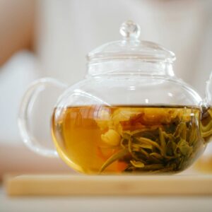 A clear glass teapot containing a brewed herbal tea blend placed on a wooden surface indoors.