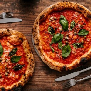 Vista superior de duas pizzas napolitanas com ingredientes frescos e talheres sobre uma mesa de madeira rústica.